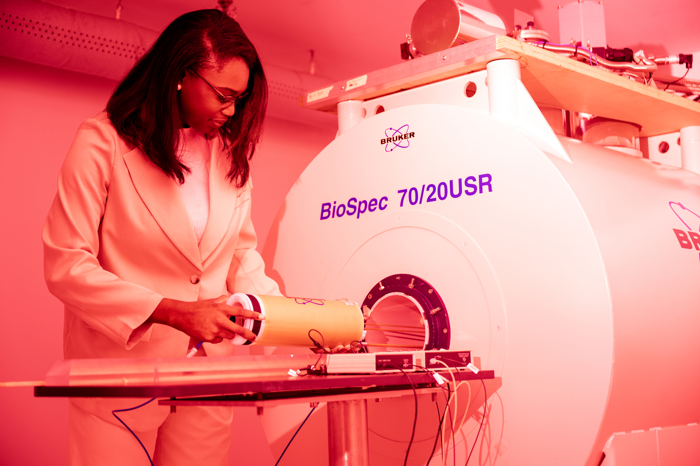 A person wearing a lab coat works with a white machine in a red-lit room.
