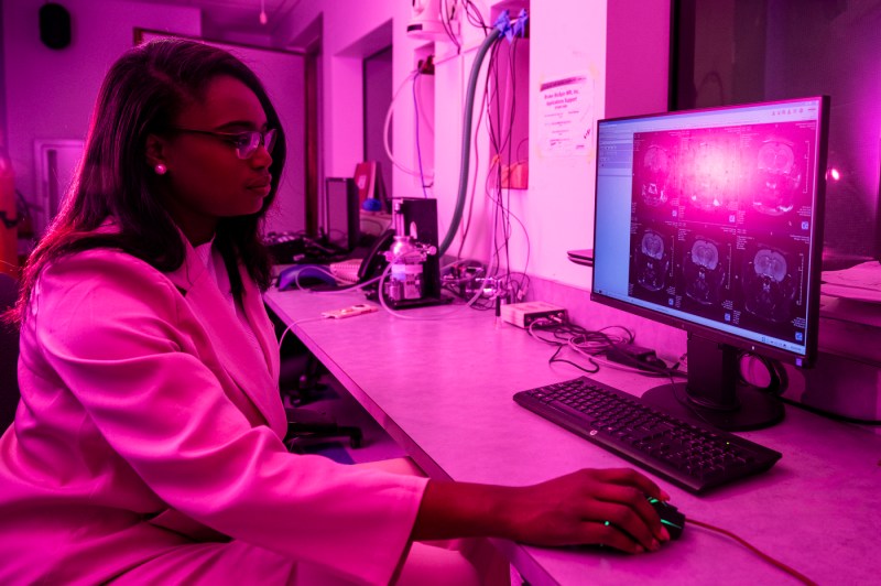 A person wearing a lab coat works with a computer in a pink-lit room. 