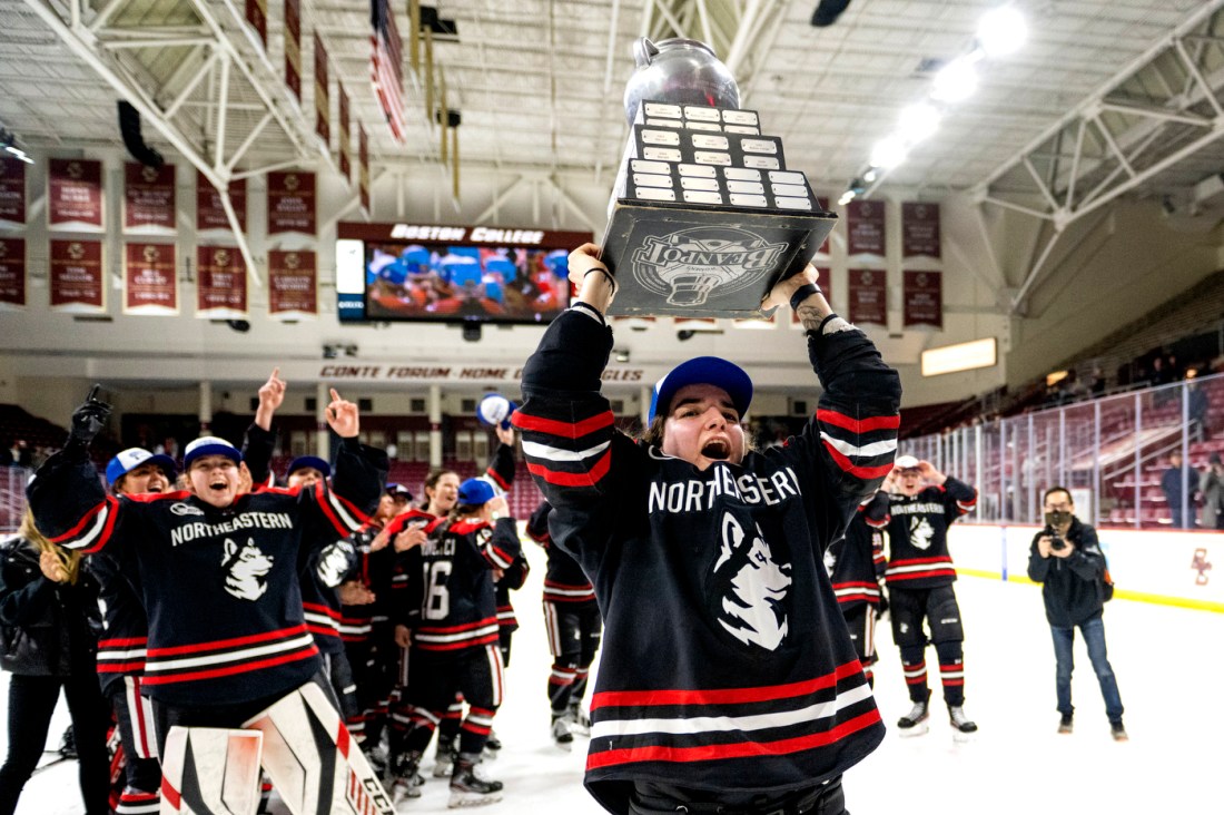 Northeastern womens hockey player lifting the beanpot trophy above her head and cheering.