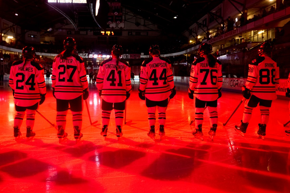 Northeastern womens hockey players lined up on the ice under red lighting, photographed from behind. 