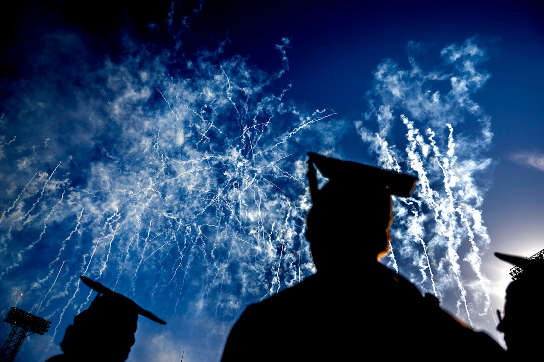 Silhouette of three Northeastern community graduates wearing caps and gowns while fireworks go off in the sky behind them. 