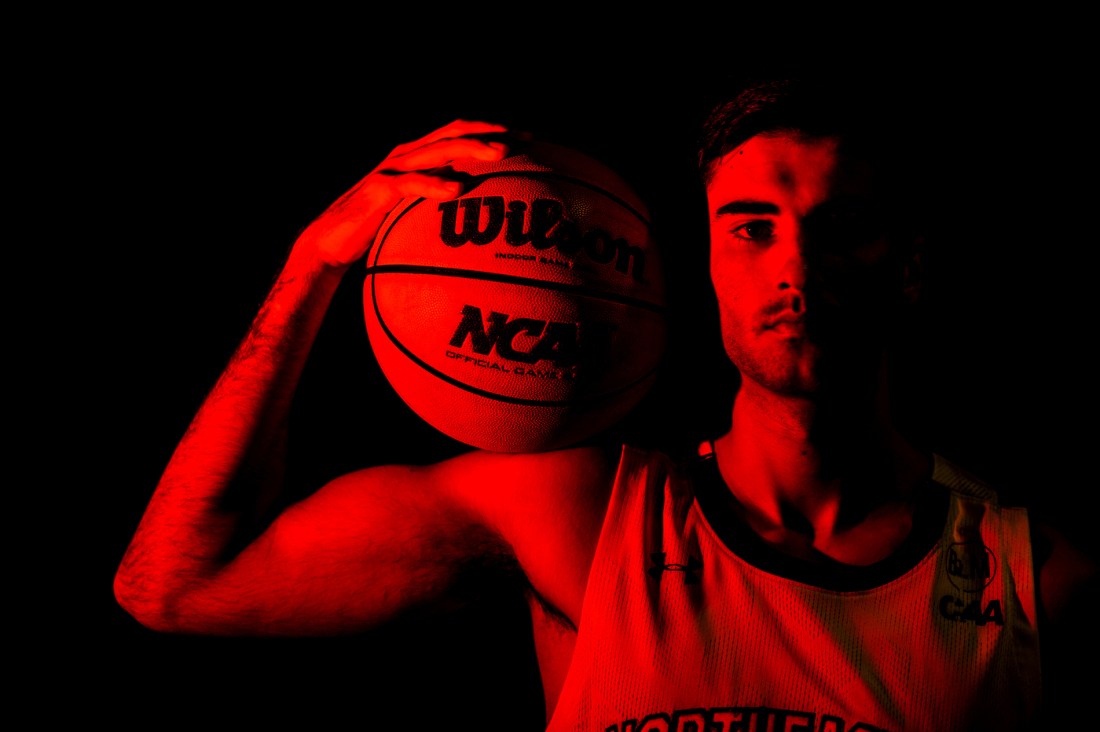 Northeastern mens basketball player standing in red lighting holding a basketball on their shoulder.