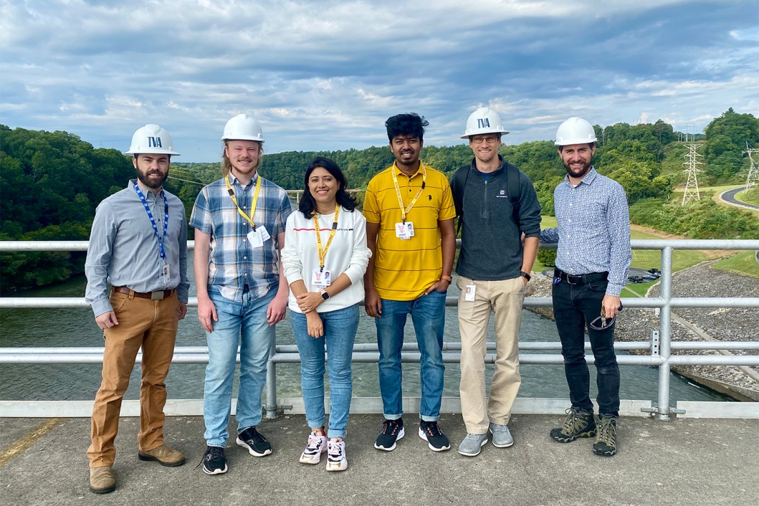 A group of six scientists and doctoral students on top of a dam in Tennessee, four wearing hard hats. 