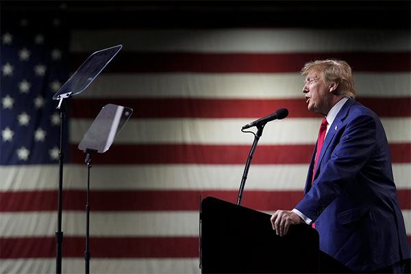 Donald Trump speaking at a microphone during a rally in front of a large United States flag.