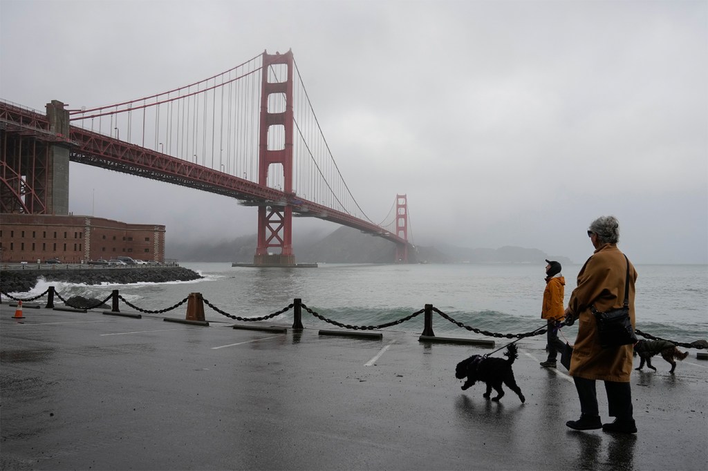 People walk their dogs at the base of the Golden Gate bridge on a foggy, overcast day. The water looks turbulent.