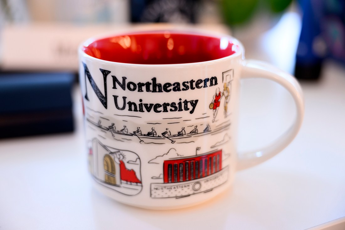 A white mug with Northeastern University written on it and doodles of student life. The inside of the mug is read.