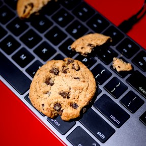 A chocolate chip cookie lays on top of a keyboard on a red-colored surface.