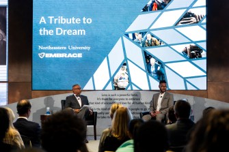 Two people sit next to each other on a stage in front of a large PowerPoint slide titled "A Tribute to the Dream."
