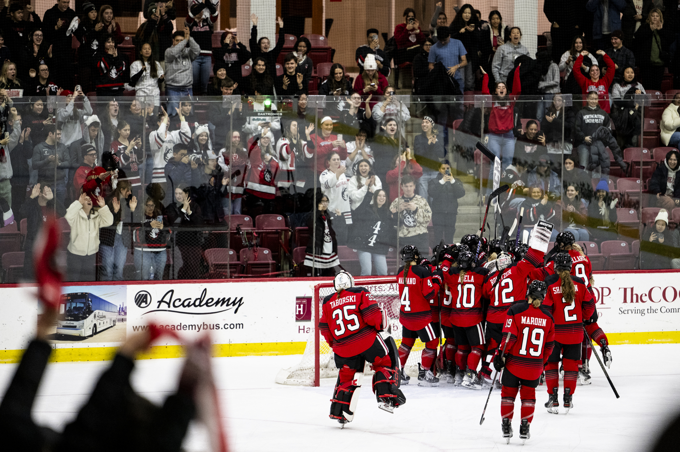 Northeastern hockey players cheer together in a circle while standing in an ice rink surrounded by cheering fans.