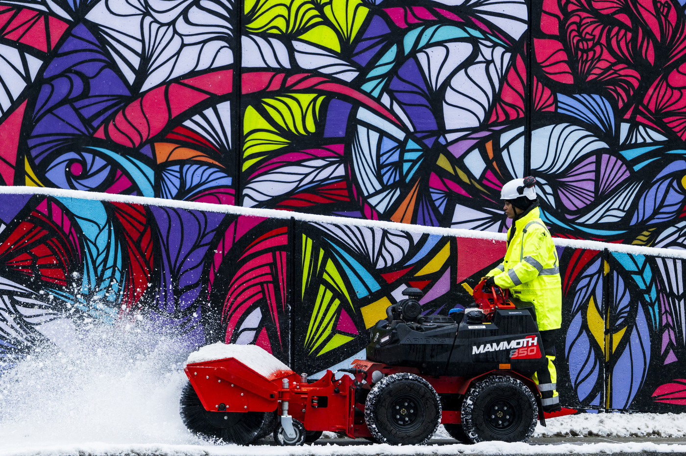 A person wearing a neon-yellow jacket uses a snowplow to clear a path in front of a multi-colored mural.