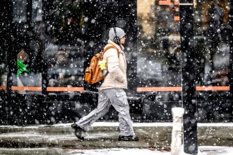 A person wearing a tan jacket, grey pants, and a burnt orange-colored backpack walks through a snowstorm.