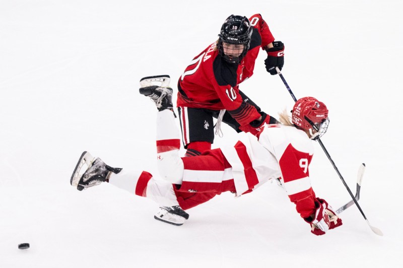 A BU hockey player falling right in front of a Northeastern hockey player on the ice.