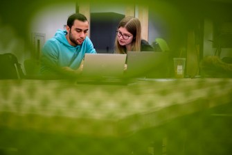 Two people, surrounded by a mesh green frame, work together at a desk in a brightly lit room.