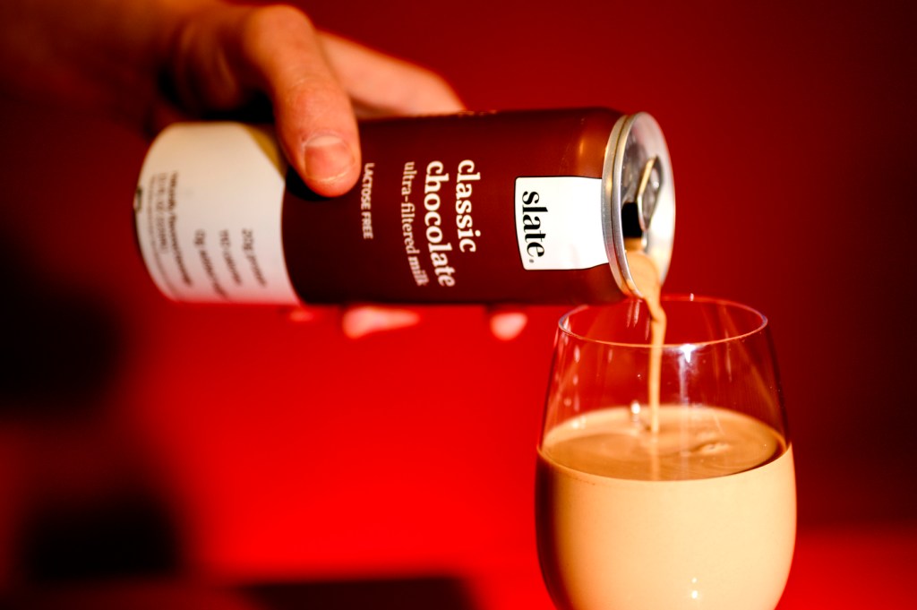Person pouring a glass of Slate Chocolate Milk into a glass in front of a red background.