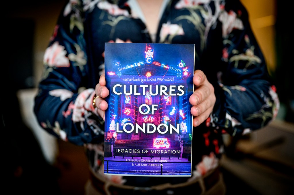 Person wearing printed shirt holding 'Cultures of London' book with blue and purple front cover.