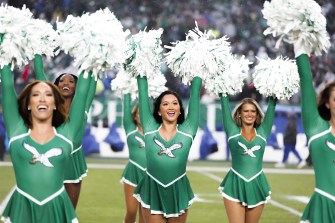 Philadelphia Eagles football team cheerleaders in green outfits holding pom poms up in the air on the football field sidelines.