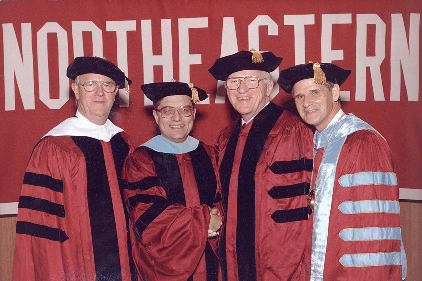 Four people, including George Matthews, wear black, red, and blue doctoral regalia while they stand in front of a red "Northeastern" sign with white text.
