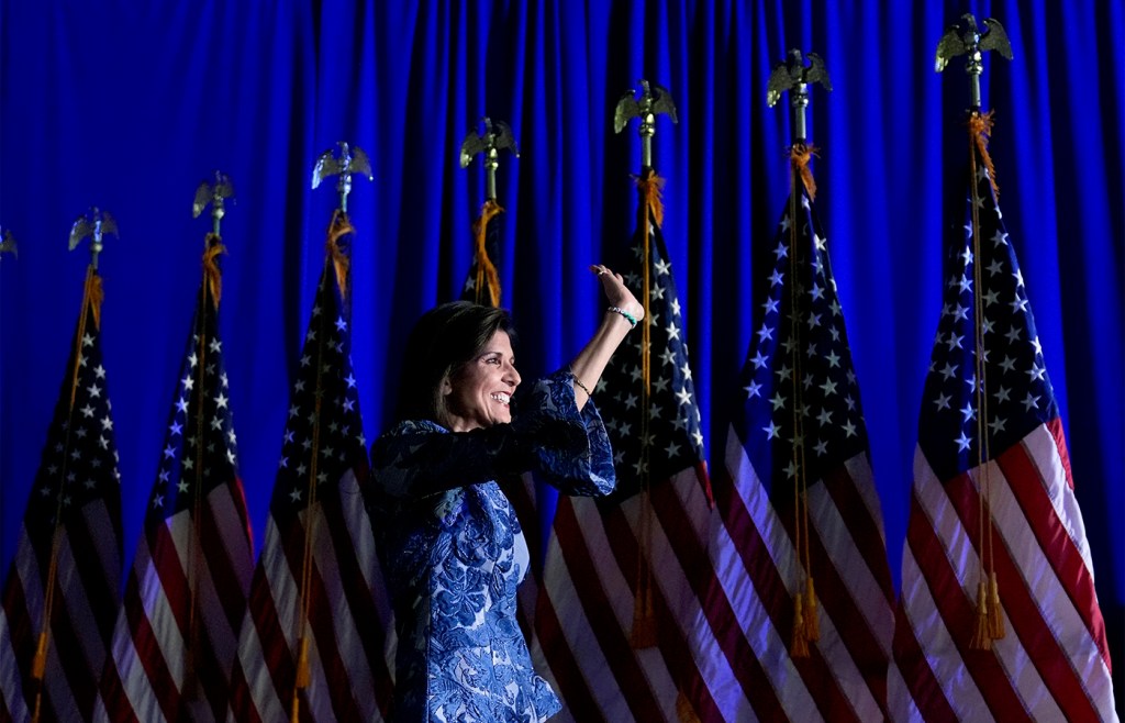 Nikki Haley standing in front of a row of US flags waving to a crowd off-camera.