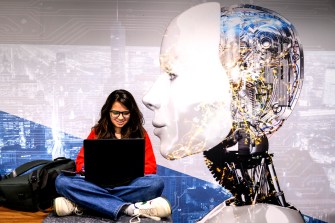 A large image of a white-colored robot looms next to a person sitting crisscrossed on a chair while they work on their laptop.