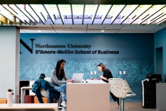 Two people work at a desk in an office in front of a blue-colored wall with black text: "Northeastern University, D'Amore-McKim School of Business."