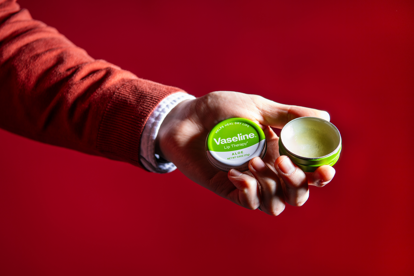 A person holding a tin of Vaseline lip balm in their hand in front of a red background.