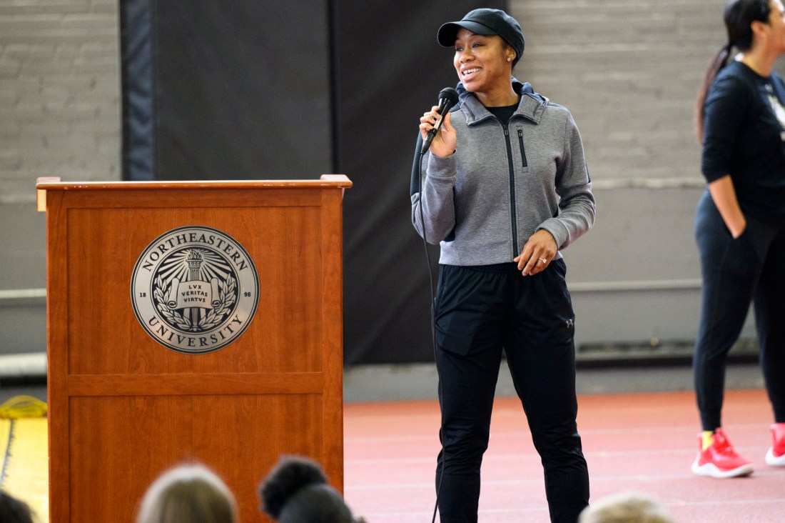A person wearing black track pants, a grey zip up, and a black baseball cap talking into a microphone.
