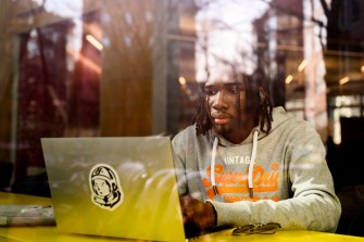 A person wearing a grey hoodie works on their laptop behind a window.