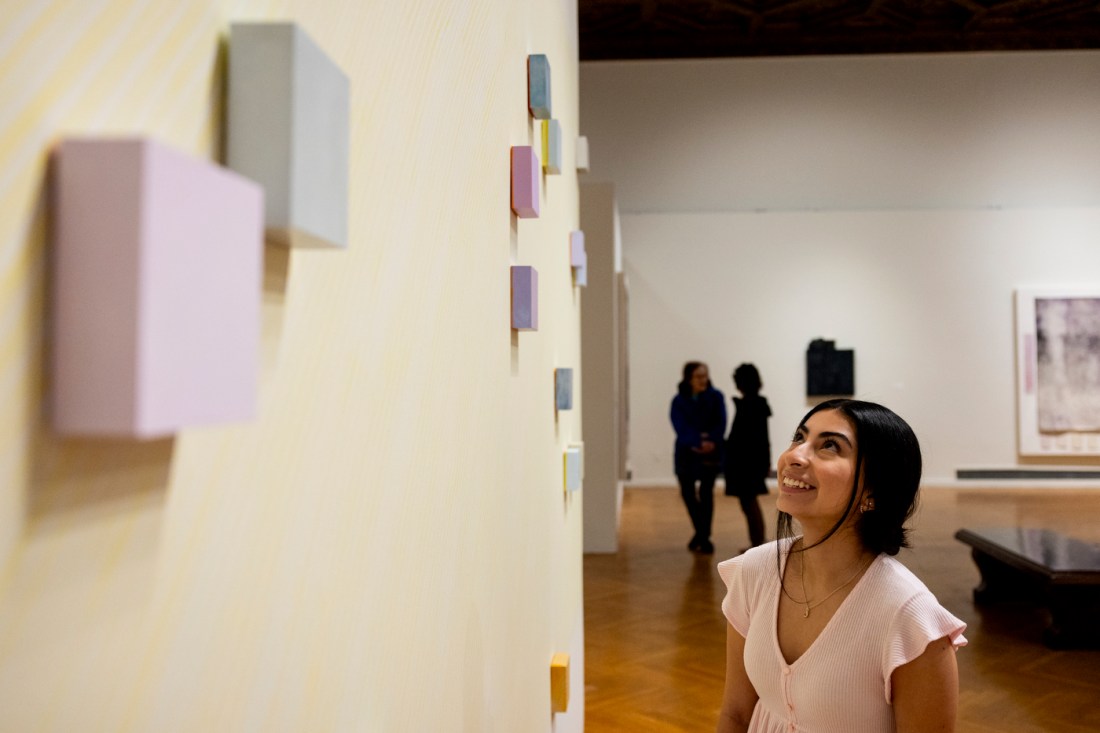 A woman looks at geometric, pastel-colored 3D shapes on a beige wall. 