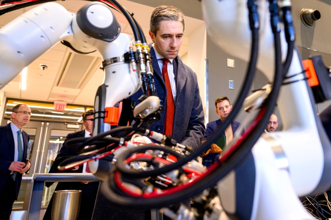 Multiple people in suits look at a robot covered in red and black wires.