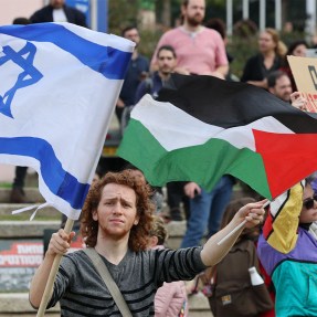 A person in a crowd holds an Israeli flag in one hand an a Palestinian flag in the other hand.