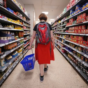 A shopper wearing a backpack carrying a blue basket through the aisles of a supermarket.