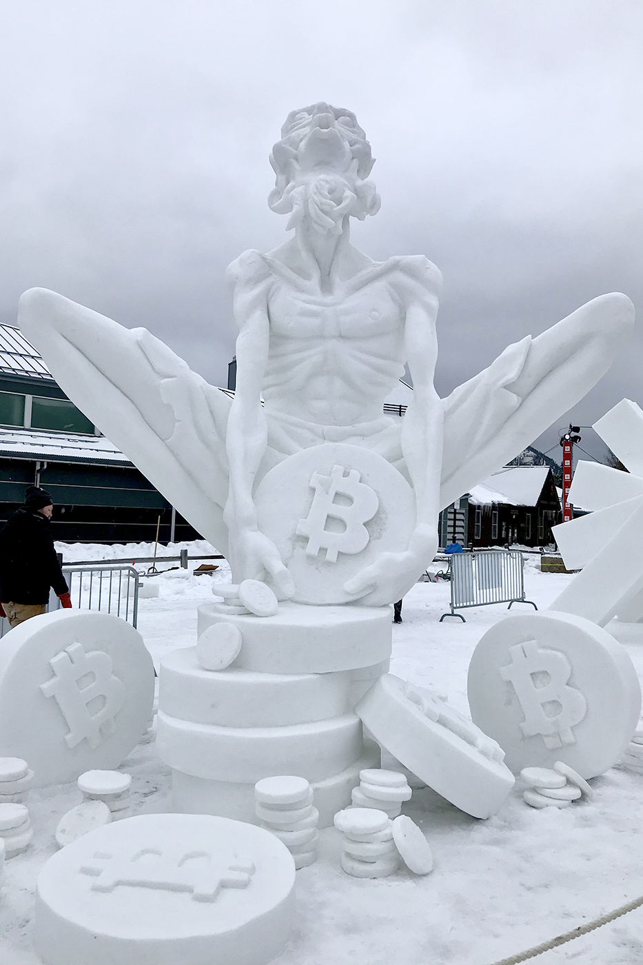 A snow sculpture resembling a figure clutching a coin emblazoned with the letter 'B' on its face.