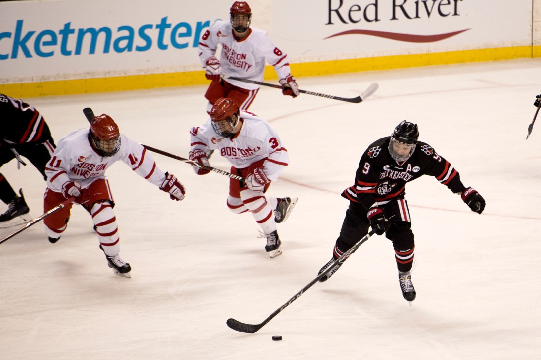 A Northeastern hockey player skates down the ice with the puck while three Boston University players chase after him.
