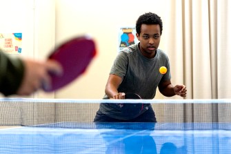 A person wearing a grey shirt and black pants plays table tennis against an opponent.