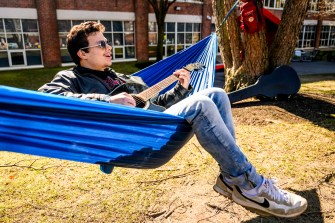 A person playing a guitar sits on a blue hammock outside on a sunny day.