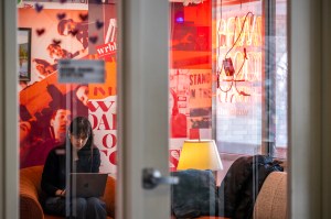 A person works on a laptop while sitting on a couch in front of a red, orange-multi colored wall.