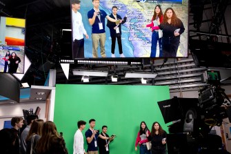 Multiple people stand in front of a green screen inside a news station studio.