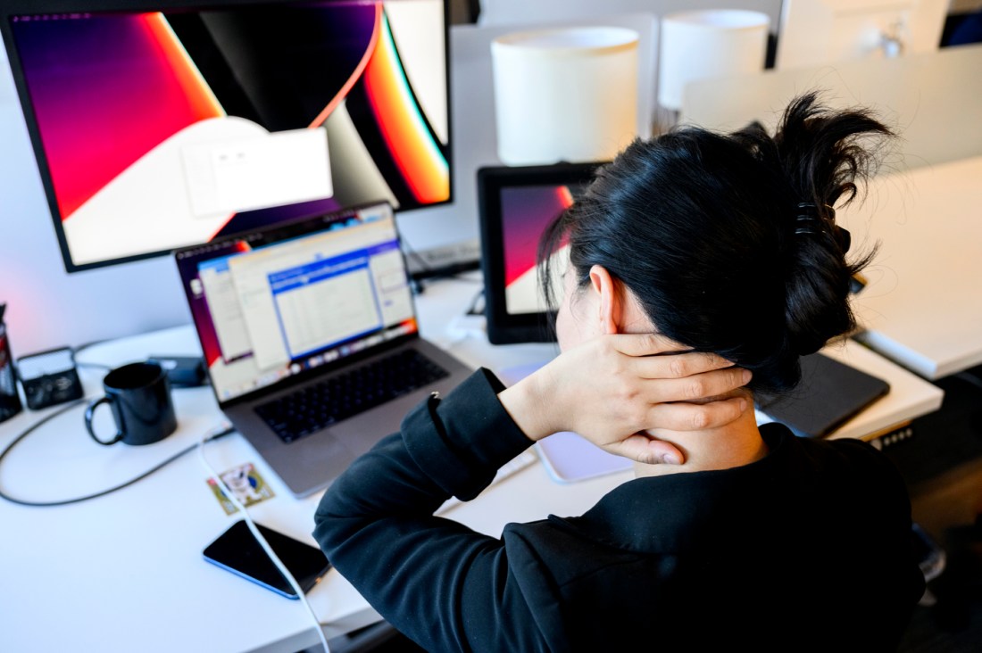 A person sitting at a desk in front of a laptop and dual monitors holding the back of their neck.