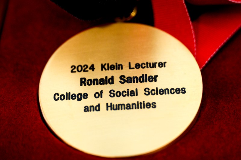 A gold medal engraved with '2024 Klein Lecturer Ronald Sandler College of Social Sciences and Humanities'. 