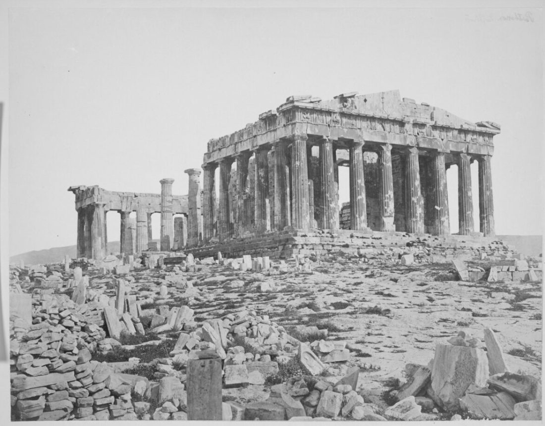 A black-and-white photograph of a ruined Greek temple, with some columns still standing and part of a pointed roof.