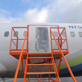 A Boeing airplane with an open area where a door might be. The area is covered with plastic and there are orange steps leading up to it.