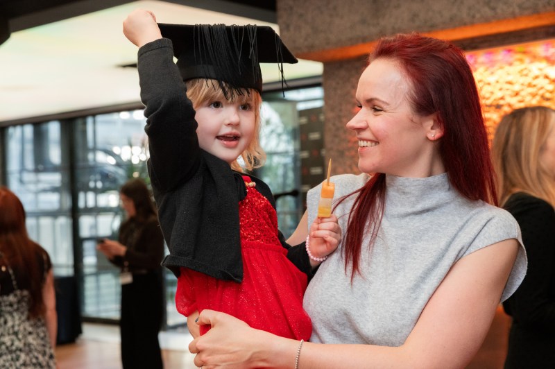 Woman holding a child wearing a graduation cap and holding a skewer with fruit on it.
