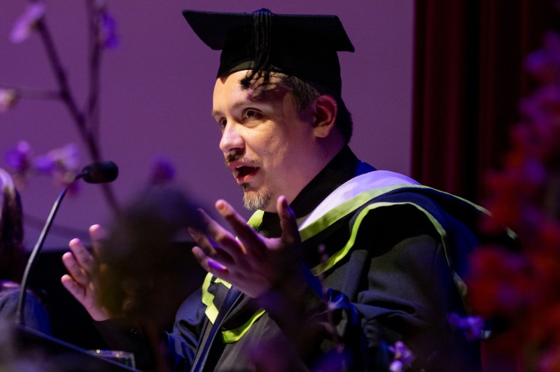 Man wearing cap and gown speaking into microphone.