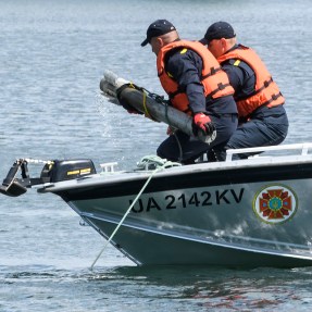 Two people in a boat wearing life jackets remove an unexploded ordnance from a lake.