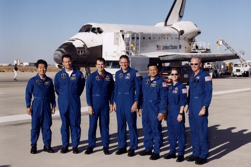 Seven member crew standing outside of a space shuttle on a runway.