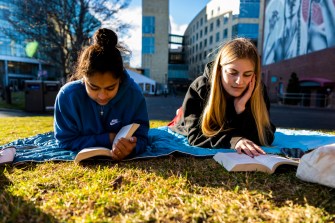 Two students reading books on a blanket in the grass.