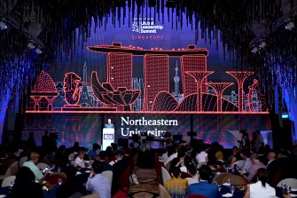 A speaker at a podium in a crowded auditorium at the Global Leadership Summit in Singapore. behind them lit up in red are famous buildings from Singapore.