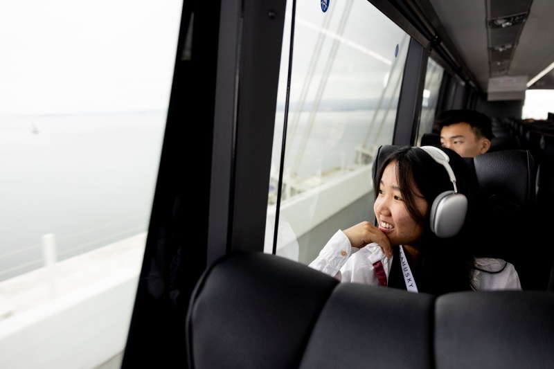 A person wearing silver headphones looks out the window to a cloudy day while sitting on a bus.