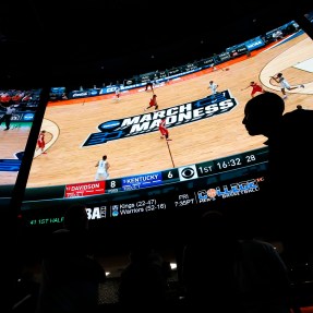 The silhouette of a person in front of a large screen airing a basketball game.