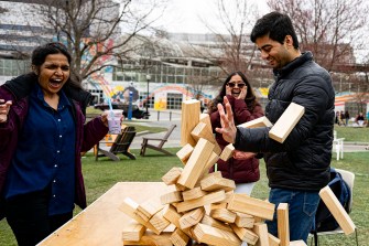 Three people react to large Jenga pieces falling off a table outside on a cloudy day.
